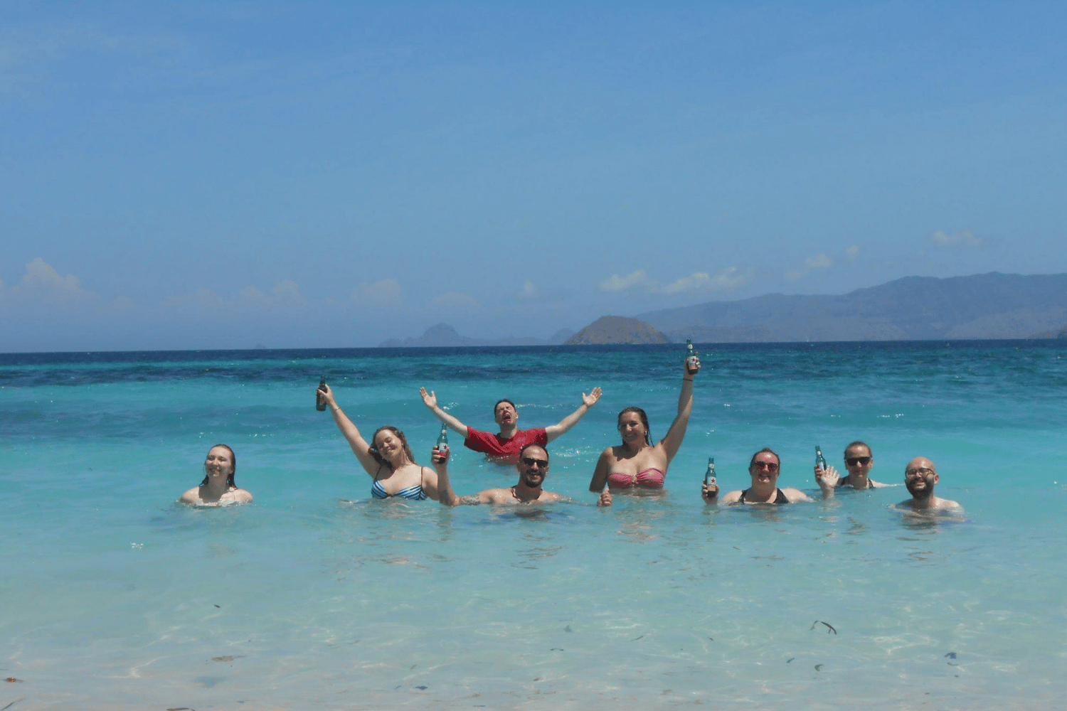 Group of people wading in clear turquoise sea, several holding bottles and raising their arms in Komodo Pink Beach