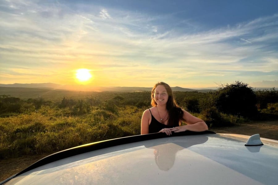 Person leaning on a car roof smiling at sunset over grassy hills in South Africa